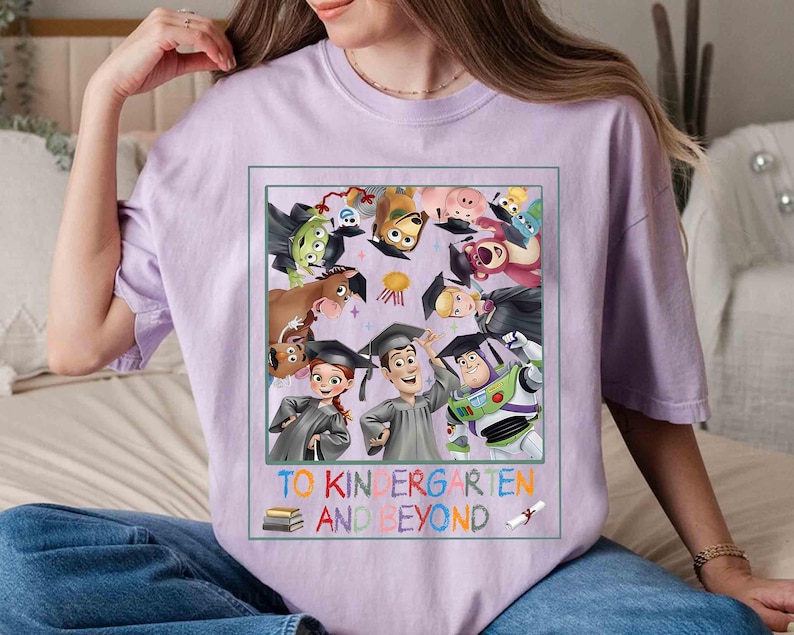 May include: A light purple t-shirt featuring a colorful graphic of Toy Story characters wearing graduation caps. The text "TO KINDERGARTEN AND BEYOND" is printed below the graphic. The shirt is a comfortable, casual style.