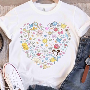 May include: White t-shirt featuring a heart-shaped design filled with colorful illustrations of fairy tale elements. Includes a crown, a magic lamp, a glass slipper, and a rose. The shirt is displayed with jeans and white sneakers.