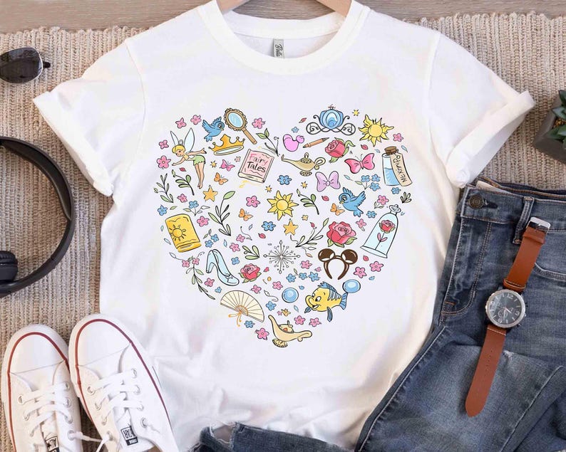 May include: White t-shirt featuring a heart-shaped design filled with colorful illustrations of fairy tale elements. Includes a crown, a magic lamp, a glass slipper, and a rose. The shirt is displayed with jeans and white sneakers.