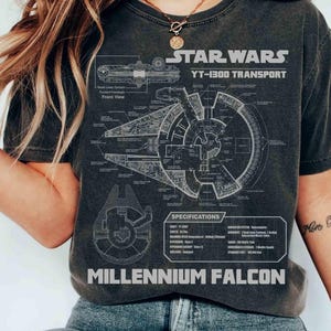 Può includere: T-shirt grigio scuro con uno schema dettagliato del Millennium Falcon, con il testo "STAR WARS YT-1300 TRANSPORT" e "MILLENNIUM FALCON". Il design include specifiche e dettagli della vista frontale.