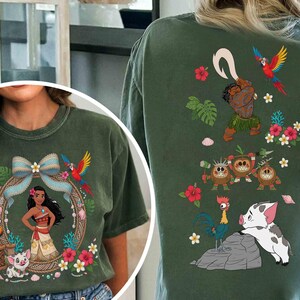 May include: A dark olive green t-shirt featuring a front graphic of a female character with long dark hair, a blue bow, a parrot, and a piglet. The back graphic displays a male character with a hook, three coconut creatures, a rooster, and a piglet.