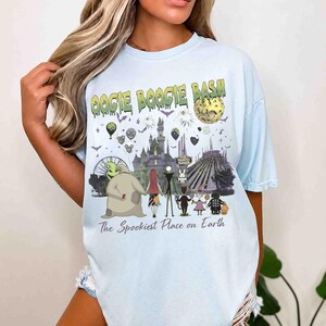 Puede incluir: Una camiseta azul claro con un estampado gr&aacute;fico que incluye el texto OOGIE BOOGIE BASH y The Spookiest Place on Earth. El dise&ntilde;o muestra varios personajes animados, un castillo, una luna, fuegos artificiales y globos festivos.