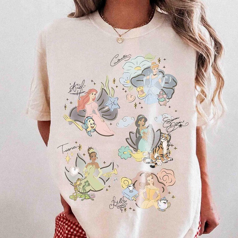 May include: A cream-colored short-sleeve t-shirt featuring a graphic print of various animated characters including Ariel, Cinderella, Jasmine, Tiana, and Belle. Each character is accompanied by their name in script and small thematic icons.
