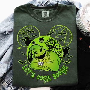 May include: Dark green t-shirt with a neon green graphic featuring a character from The Nightmare Before Christmas. The design includes bats, spiderwebs, and the text "Let's Oogie Boogie". The shirt is laid on a denim jacket and black pants.