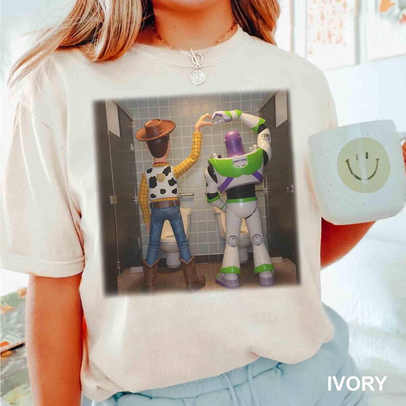 May include: An off-white short-sleeve t-shirt featuring a graphic print of Woody and Buzz Lightyear standing in bathroom stalls, forming a heart shape with their hands. The shirt is paired with a light-colored mug with a yellow smiley face.