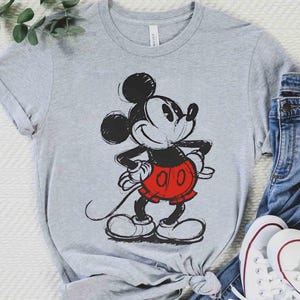 May include: A gray t-shirt featuring a classic Mickey Mouse graphic. The cartoon character is depicted in black, red, and white, with a sketched design. The shirt is paired with blue jeans and white sneakers.