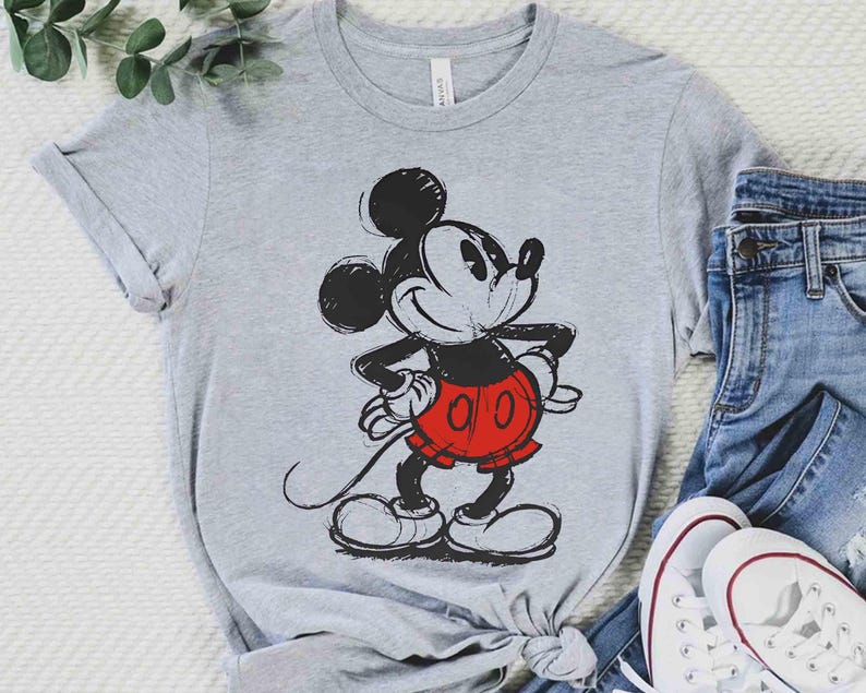 May include: A gray t-shirt featuring a classic Mickey Mouse graphic. The cartoon character is depicted in black, red, and white, with a sketched design. The shirt is paired with blue jeans and white sneakers.