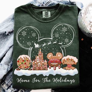 May include: Dark green t-shirt with a white graphic of Mickey Mouse ears filled with snowflakes and a sleigh. Below, gingerbread castle and cookies are on a snow-covered base. The text "Home For The Holidays" is at the bottom.