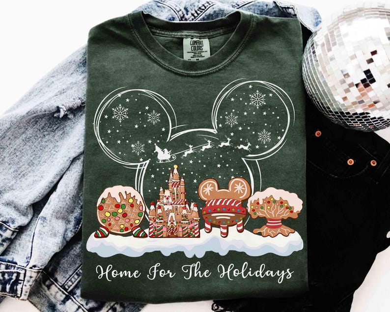 May include: Dark green t-shirt with a white graphic of Mickey Mouse ears filled with snowflakes and a sleigh. Below, gingerbread castle and cookies are on a snow-covered base. The text "Home For The Holidays" is at the bottom.