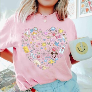 May include: Pink t-shirt featuring a heart-shaped design filled with colorful illustrations of fairy tale elements, including a magic lamp, a glass slipper, and a rose. The shirt also has the text "Fairy Tales".