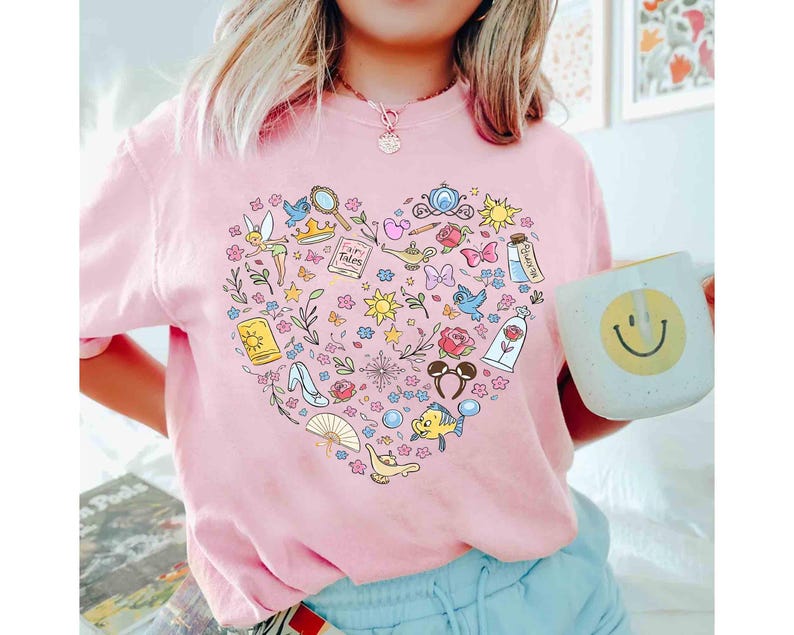 May include: Pink t-shirt featuring a heart-shaped design filled with colorful illustrations of fairy tale elements, including a magic lamp, a glass slipper, and a rose. The shirt also has the text "Fairy Tales".