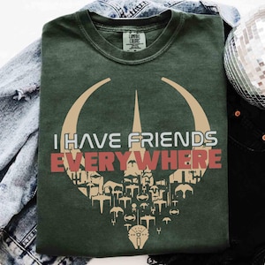 以下が含まれることがあります： 「I HAVE FRIENDS EVERYWHERE」の白と赤の文字が入った濃い緑色のTシャツ。様々な形や図形を使ったグラフィックデザインが特徴です。Tシャツはデニムジャケットと黒いパンツの上に置かれています。