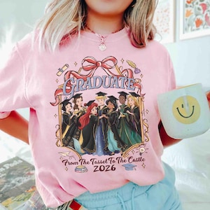 Pode incluir: Camiseta rosa claro com um gr&aacute;fico com a palavra "GRADUATE" e personagens de desenhos animados em trajes de formatura. O texto "From The Tassel To The Castle 2026" tamb&eacute;m est&aacute; inclu&iacute;do. Uma caneca branca com um rosto sorridente est&aacute; na imagem.