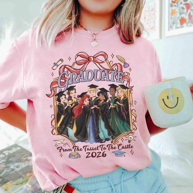 Pode incluir: Camiseta rosa claro com um gr&aacute;fico com a palavra "GRADUATE" e personagens de desenhos animados em trajes de formatura. O texto "From The Tassel To The Castle 2026" tamb&eacute;m est&aacute; inclu&iacute;do. Uma caneca branca com um rosto sorridente est&aacute; na imagem.