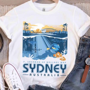 May include: White t-shirt featuring a colorful graphic of Sydney, Australia, with the Sydney Harbour Bridge, palm trees, and underwater scenes with fish. The text reads "SYDNEY AUSTRALIA" and "P. Sherman 42 Wallaby Way."