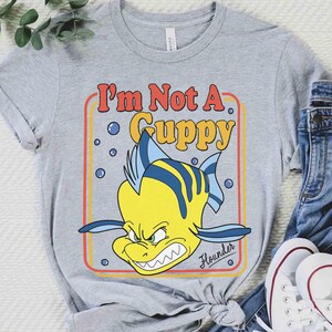 Op de afbeelding: Een grijs t-shirt met een cartoonafbeelding van Bot uit De Kleine Zeemeermin. De gele vis heeft blauwe strepen en een boze uitdrukking. De tekst "I'm Not A Guppy" staat in een rood en geel retro lettertype.