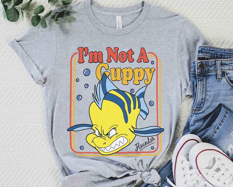 Op de afbeelding: Een grijs t-shirt met een cartoonafbeelding van Bot uit De Kleine Zeemeermin. De gele vis heeft blauwe strepen en een boze uitdrukking. De tekst "I'm Not A Guppy" staat in een rood en geel retro lettertype.