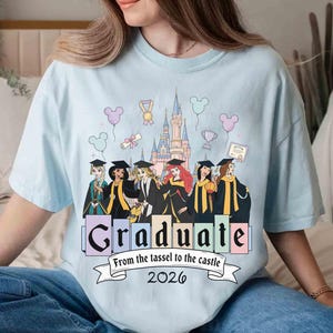 May include: Light blue t-shirt featuring a graphic of Disney princesses in graduation attire in front of a castle. The word "Graduate" is displayed with the text "From the tassel to the castle 2026" below.
