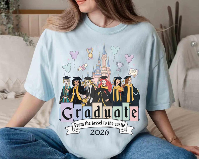 May include: Light blue t-shirt featuring a graphic of Disney princesses in graduation attire in front of a castle. The word "Graduate" is displayed with the text "From the tassel to the castle 2026" below.