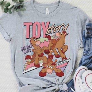 May include: A light grey t-shirt with a graphic of the Bullseye character from Toy Story. The design includes the words "TOY Story" in pink and the text "SINCE 1999". The shirt is styled with blue jeans and white trainers.