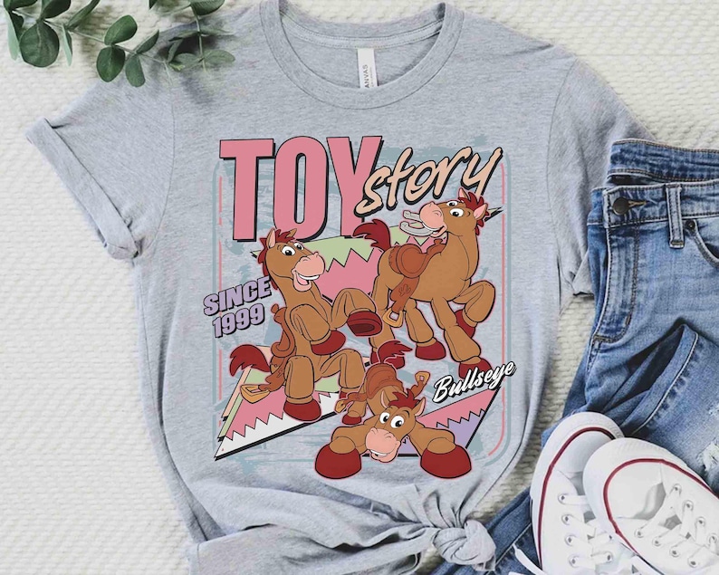 May include: A light grey t-shirt with a graphic of the Bullseye character from Toy Story. The design includes the words "TOY Story" in pink and the text "SINCE 1999". The shirt is styled with blue jeans and white trainers.