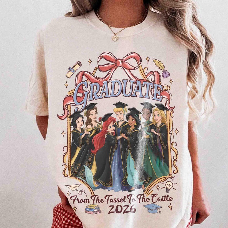 Pode incluir: Camiseta creme com um gr&aacute;fico de princesas da Disney em trajes de formatura. A palavra "GRADUATE" &eacute; exibida acima das princesas, com o texto "From The Tassel To The Castle 2026" abaixo. O design inclui um la&ccedil;o vermelho e elementos decorativos.