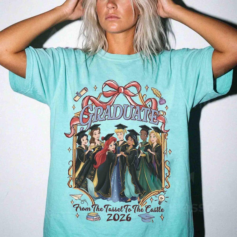 Pode incluir: Camiseta azul clara com um design gr&aacute;fico. O design inclui a palavra "GRADUATE" em uma fonte decorativa, um la&ccedil;o vermelho e ilustra&ccedil;&otilde;es de princesas da Disney em trajes de formatura. O texto "From The Tassel To The Castle 2026" tamb&eacute;m est&aacute; inclu&iacute;do.