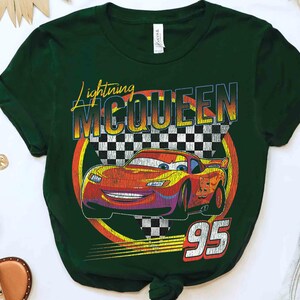 May include: Dark green t-shirt with a graphic of Lightning McQueen from the Cars film. The design includes the text "Lightning McQueen" and the number "95" with a checkered flag background. The car is red and yellow.