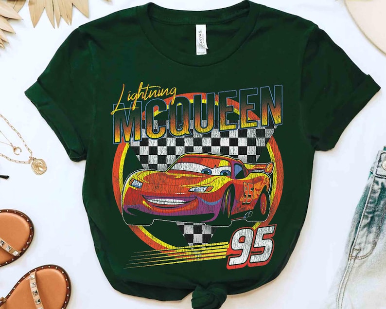 May include: Dark green t-shirt with a graphic of Lightning McQueen from the Cars film. The design includes the text "Lightning McQueen" and the number "95" with a checkered flag background. The car is red and yellow.