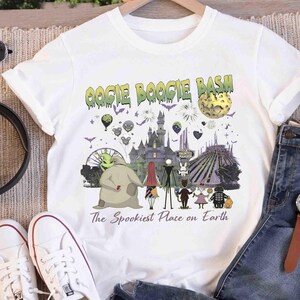 Puede incluir: Una camiseta blanca de manga corta con un estampado gr&aacute;fico que incluye el texto OOGIE BOOGIE BASH y The Spookiest Place on Earth. El dise&ntilde;o muestra un castillo, una luna, globos y varios personajes animados en un estilo oscuro.