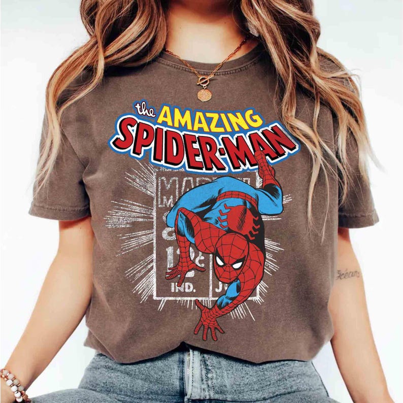 May include: A brown t-shirt with a vintage comic book graphic of Spider-Man. The design features the text "The Amazing Spider-Man" in a bold, colourful font above the character. The graphic is in a retro comic book style, with a dynamic pose.