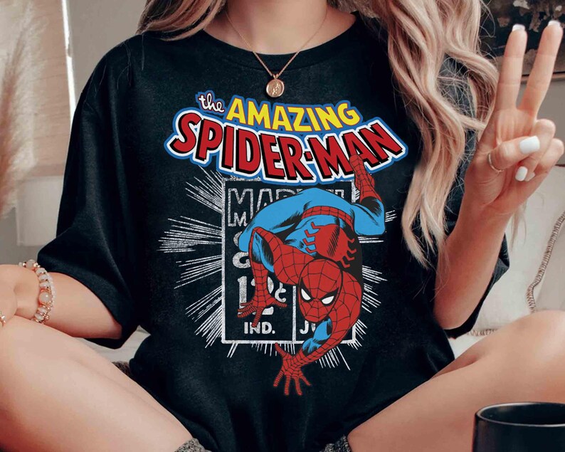 May include: Black t-shirt with a graphic of Spider-Man in a dynamic pose. The design includes the words "The Amazing Spider-Man" in red and blue, with a white outline. The shirt has a classic comic book style.