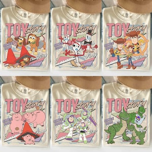 May include: Cream-coloured t-shirts featuring various Toy Story characters and the words "TOY STORY" in pink. Each shirt has a different character design, including Slinky Dog, Forky, Woody, Hamm, Buzz, and Rex. The shirts also have the text "SINCE 1995" or "SINCE 2019".
