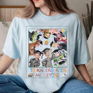 May include: Light blue t-shirt featuring a colorful graphic of animated characters wearing graduation caps. The graphic includes the text "TO KINDERGARTEN AND BEYOND" in a playful font, with a stack of books and a diploma illustration.