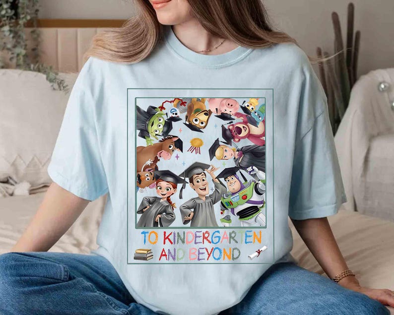 May include: Light blue t-shirt featuring a colorful graphic of animated characters wearing graduation caps. The graphic includes the text "TO KINDERGARTEN AND BEYOND" in a playful font, with a stack of books and a diploma illustration.