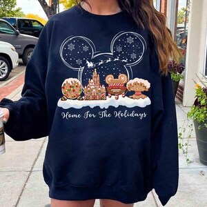 May include: Navy blue sweatshirt featuring a festive holiday design. The graphic includes a Mickey Mouse silhouette with snowflakes, a gingerbread castle, and the text "Home for the Holidays". The design incorporates gingerbread cookies and a sleigh.