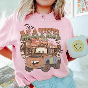 K&ouml;nnte beinhalten: Ein hellrosa T-Shirt mit einem Grafikdruck der Figur Tow Mater aus dem Film Cars. Das Design enth&auml;lt den Text Tow Mater, eine W&uuml;stenlandschaft und eine Seilumrandung. Das T-Shirt wird mit einer hellblauen Tasse mit gelbem Smiley-Gesicht kombiniert.