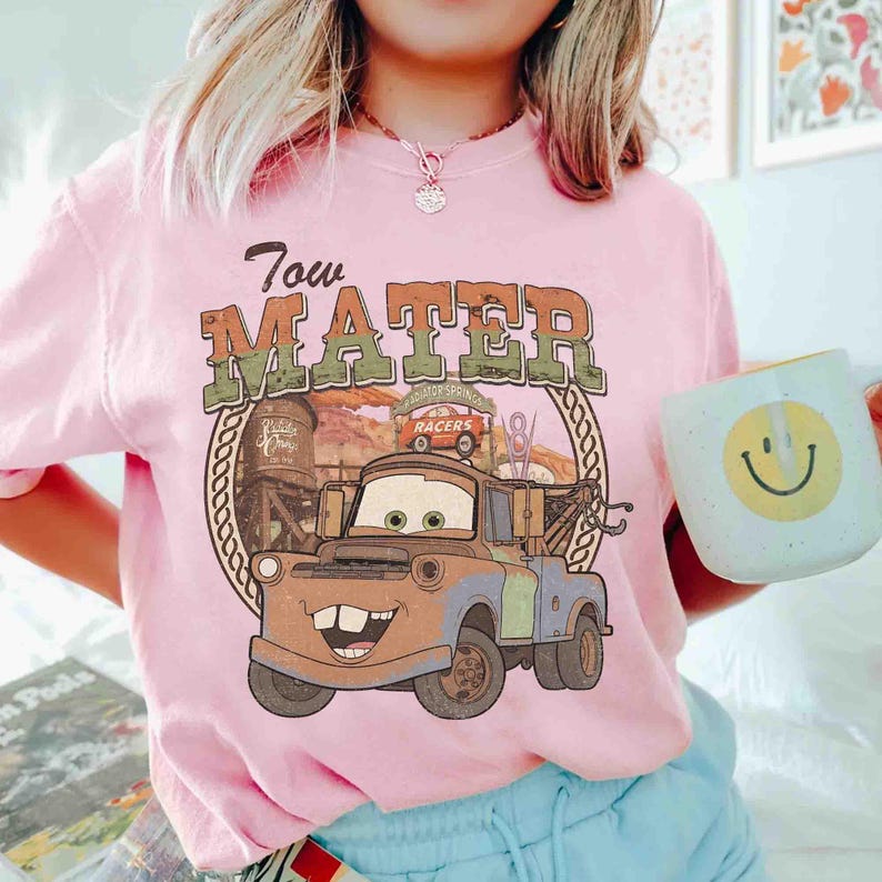 K&ouml;nnte beinhalten: Ein hellrosa T-Shirt mit einem Grafikdruck der Figur Tow Mater aus dem Film Cars. Das Design enth&auml;lt den Text Tow Mater, eine W&uuml;stenlandschaft und eine Seilumrandung. Das T-Shirt wird mit einer hellblauen Tasse mit gelbem Smiley-Gesicht kombiniert.