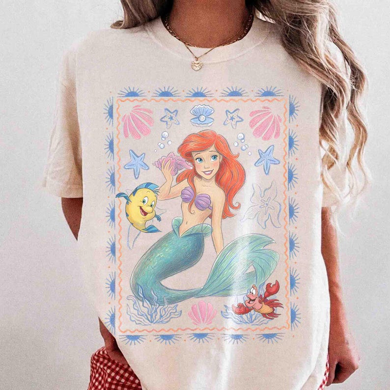 Op de afbeelding: Een cr&egrave;mekleurig t-shirt met een rechthoekige grafische print van een roodharige zeemeermin met een groenblauwe staart, een gele vis en een kleine rode krab. Het ontwerp is omringd door een blauwe rand met zonnestralen en schelpen.
