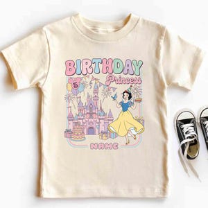 May include: A cream-coloured t-shirt with a colourful graphic featuring the text "BIRTHDAY Princess" and a cartoon princess. The graphic includes a castle, balloons, and a cake. Black canvas trainers with white laces are next to the shirt.