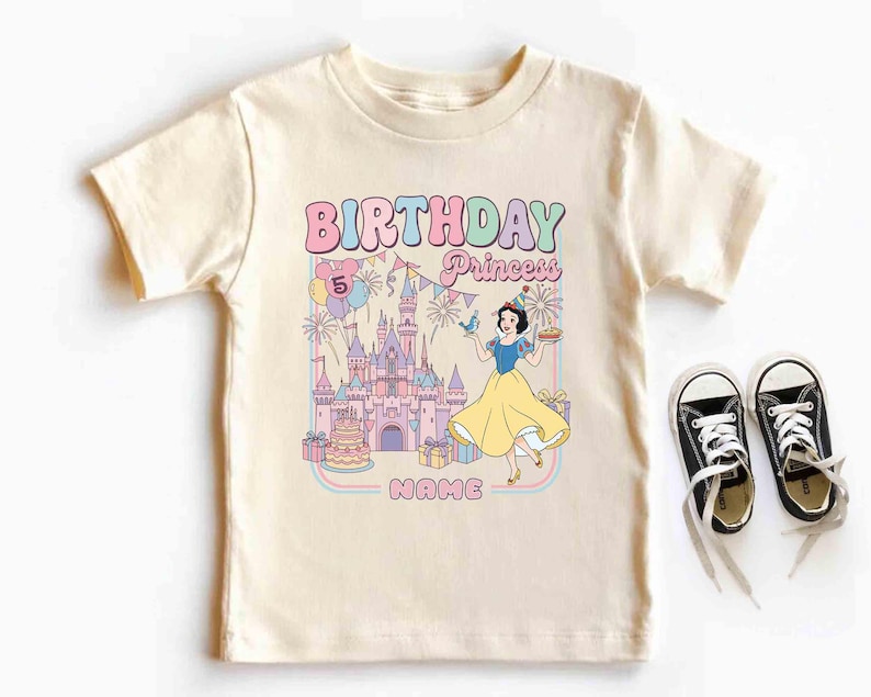 May include: A cream-coloured t-shirt with a colourful graphic featuring the text "BIRTHDAY Princess" and a cartoon princess. The graphic includes a castle, balloons, and a cake. Black canvas trainers with white laces are next to the shirt.