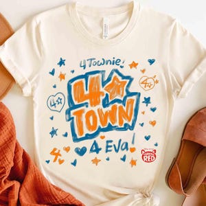Può includere: T-shirt color crema con le parole "4 Town" in arancione e blu, circondate da stelle e cuori. La maglietta presenta anche il testo "4 Eva!" e un piccolo logo "Turning Red". La maglietta è abbinata a sandali marroni e una sciarpa arancione.