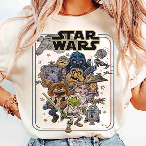 May include: A cream-colored t-shirt featuring a graphic print of Star Wars characters reimagined as Muppets. The design includes Kermit the Frog as Luke Skywalker, Miss Piggy as Princess Leia, and Fozzie Bear as Han Solo, with the Star Wars logo above.