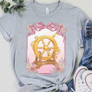 Puede incluir: Una camiseta gris con un dise&ntilde;o de tim&oacute;n dorado, detalles rosas y florales. El gr&aacute;fico est&aacute; enmarcado por una cinta rosa y flores. La camiseta se combina con vaqueros azules y zapatillas blancas.