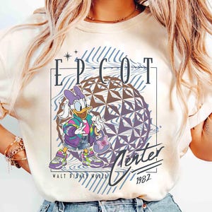 May include: Cream-colored t-shirt featuring Daisy Duck in a colorful outfit, standing in front of a geometric sphere. The shirt has the text "EPCOT Center" and "Winter 1982" with the Walt Disney World logo.
