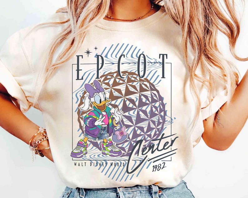 May include: Cream-colored t-shirt featuring Daisy Duck in a colorful outfit, standing in front of a geometric sphere. The shirt has the text "EPCOT Center" and "Winter 1982" with the Walt Disney World logo.