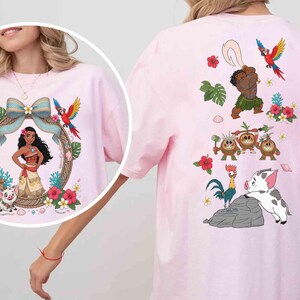 May include: A light pink t-shirt featuring a circular graphic of a female character with long dark hair, a blue bow, a parrot, and a pig. The back of the shirt displays a larger print of the same character, a parrot, three coconut creatures, a rooster, and a pig.