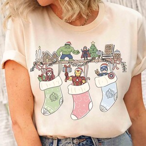 Puede incluir: Camiseta color crema con un diseño festivo de Marvel. El diseño incluye calcetines navideños con personajes de los Vengadores, una repisa decorada y un árbol de Navidad. Es una prenda informal con temática navideña.