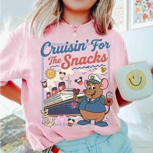 Pode incluir: Camiseta rosa com o texto "Cruisin' For the Snacks" e um desenho animado de um capit&atilde;o rato em um cruzeiro. O rato est&aacute; segurando uma bebida e sorvete. Uma caneca com um rosto sorridente &eacute; segurada na m&atilde;o.