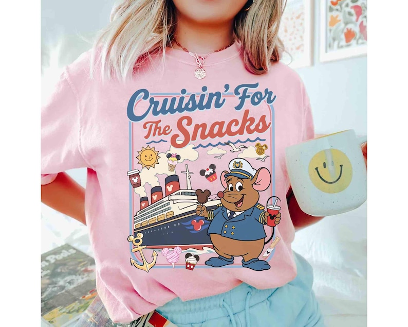 Pode incluir: Camiseta rosa com o texto "Cruisin' For the Snacks" e um desenho animado de um capit&atilde;o rato em um cruzeiro. O rato est&aacute; segurando uma bebida e sorvete. Uma caneca com um rosto sorridente &eacute; segurada na m&atilde;o.